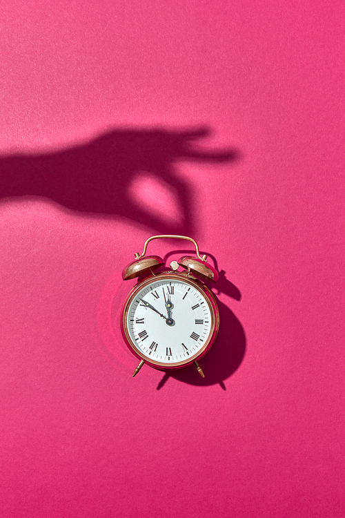 Retro alarm clock painted golden with hard shadows from woman's hand holding it on a hot pink background, copy space.