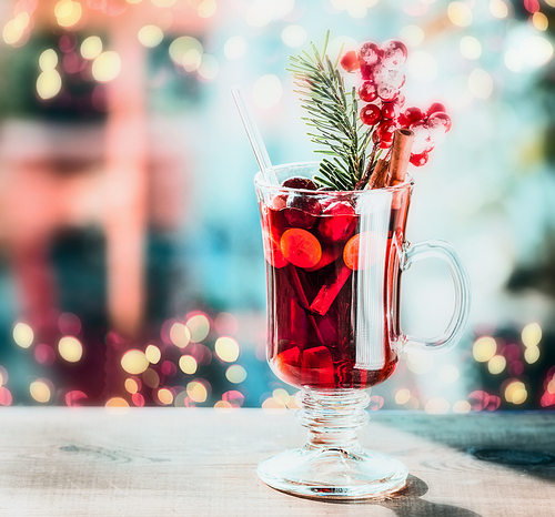 Glass of spiced mulled wine with berries and fir branch on table at festive bokeh lighting background