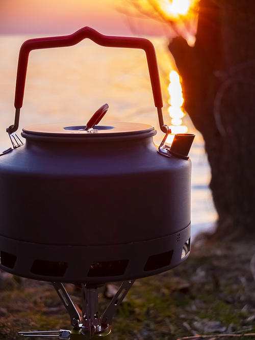 Boiling water in a touristic kettle outdoors next to the water at sunset.