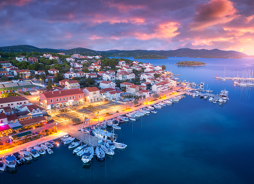 Aerial view of boats and yachts in port and city at night. Summer landscape with city lights, buildings, illuminated streets, mountain, motorboats, blue sea, colorful sky at sunset. Top view. Croatia