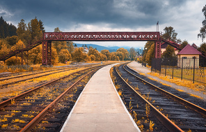 Rural railway station in autumn in cloudy day. Industrial landscape with old railway platform, orange trees, bridge, building, cloudy sky. Railroad and beautiful forest in countryside. Railway in fall