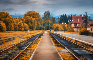 Rural railway station in autumn in cloudy day. Industrial landscape with old railway platform, orange trees, buildings, overcast sky. Railroad and beautiful forest in countryside. Railway in fall