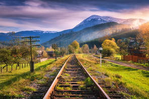 Railroad in mountains with snowy peaks at sunset in autumn. Industrial landscape with railway station, orange trees, green grass, buildings, rocks, purple sky with clouds in fall. Railway platform