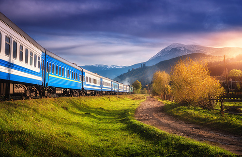 Moving train in mountains at sunset in autumn. Industrial landscape with passenger speed train on railroad, dirt road, snowy rocks, orange trees, green grass, purple sky in fall. Rural railway station