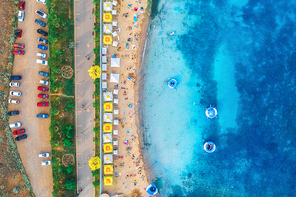 Aerial view of sandy beach with colorful umbrellas, swimming people in sea with transparent blue water, swim rings, green trees and cars on the promenade at sunset in summer. Vacations. Top view