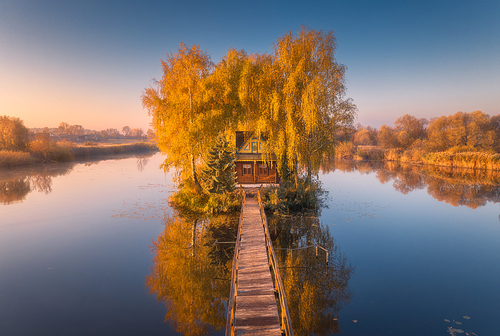 Old fisherman house and wooden pier at sunrise in autumn. Aerial view. Beautiful landscape with house on small island on the lake, colorful trees, jetty, reflection in water. Fall in Ukraine. Top view