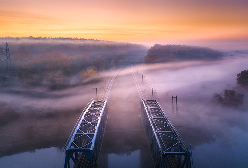 Aerial view of railroad bridge in fog at sunrise in autumn. Colorful landscape with foggy trees, fields, mist, river, railway station, orange sky. Top view. Railroad in fog in dusk in fall. Industrial