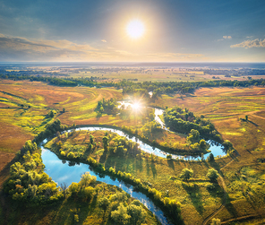 Aerial view of beautiful curving river at sunrise in summer. View from air. Turns of river, green meadows, grass and trees, blue sky, sun at dawn. Colorful aerial landscape of river coast. Top view