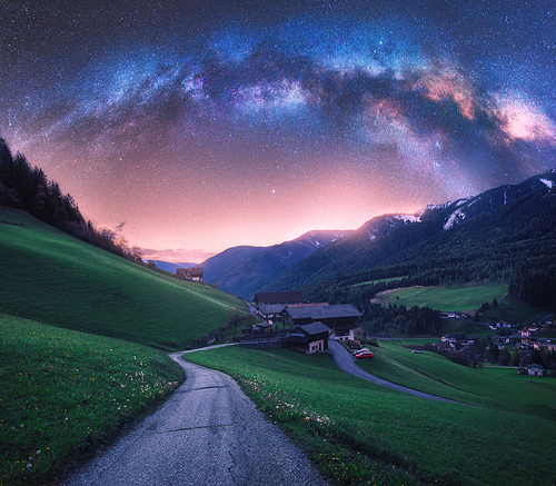 Arched Milky Way over the rural mountain road in summer in Italy. Beautiful night landscape with starry sky, milky way arch, winding road in mountain village, hills, green meadows and buildings. Space