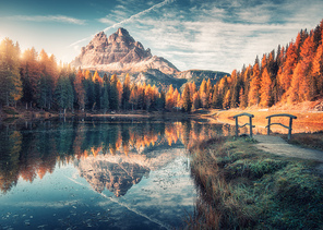 Lake with reflection in mountains at sunrise in autumn in Dolomites, Italy. Landscape with Antorno lake, small wooden bridge, trees with orange leaves, high rocks, blue sky in fall. Colorful forest