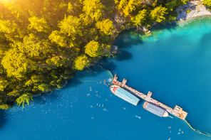 Aerial view of boats near wooden jetty in blue see and green trees at sunset in summer. Colorful landscape with swimming people in lake with clear water, boat, yacht and mountain forest. Top view