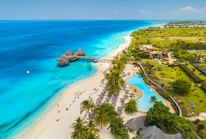 Aerial view of beautiful hotel on the sea, pool, umbrellas and green palm trees on the sandy beach at sunny summer day. Top view. Tropical landscape with wooden hotel on the blue water, trees, boats