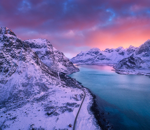 Aerial view of beautiful snowy mountains, sea, road, sky with pink clouds at sunset in winter. Lofoten islands, Norway. Top view of road, rocks in snow, sea coast, azure water. View from above. Nature