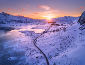 Aerial view of winding road, snow covered mountains, orange sky at sunset in winter. Landscape with sea, frosty coast, snowy rocks, roadway. Lofoten islands, Norway. Frozen sea shore. View from above
