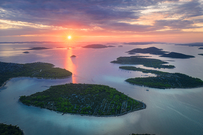 Aerial view of beautiful small islands in Adriatic sea at sunset in summer in Croatia. Top view of blue water, green trees on the mountains, colorful sky with clouds and orange sun. Tropical landscape