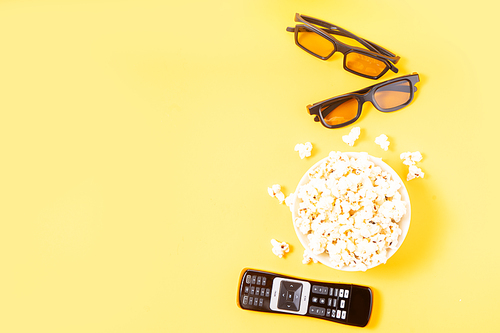 bowl of popcorn, nd remote control for TV and 3d glasses over yellow background with copy space, movie and cinema concept