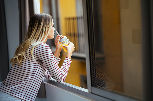 Young blonde woman drinking a glass of natural orange juice, leaning out the window of her home.
