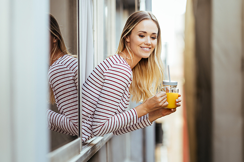 Smiling blonde woman drinking a glass of natural orange juice, leaning out the window of her home.