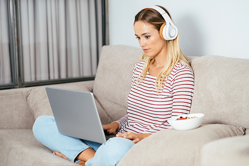 Young blonde woman with headphones and laptop on the sofa at home.