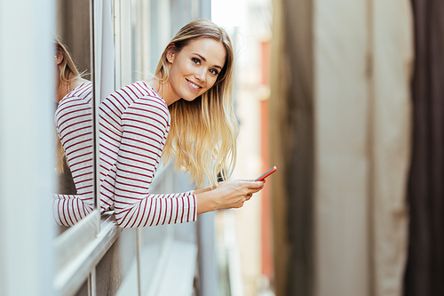 Young blonde woman using a smartphone . Girl leaning out of her house window.