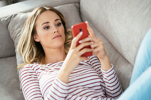 Young blonde woman using her smartphone lying on the sofa at home.