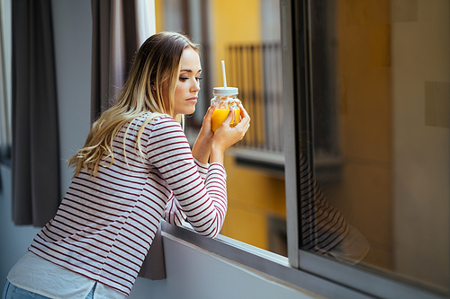Young caucasian woman drinking a glass of natural orange juice, leaning out the window of her home.