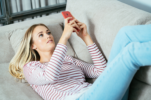 Young caucasian woman using her smartphone lying on the sofa at home.