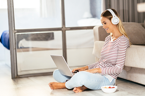 Smiling young woman with headphones and laptop on the sofa at home.