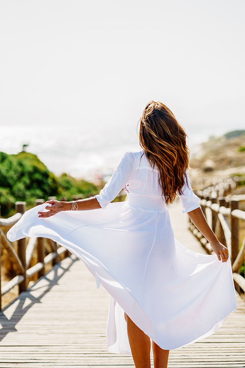 Rear view of a young woman wearing a beautiful white dress in Spanish fashion on a boardwalk on the beach.