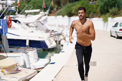 Young man with an athletic body running shirtless through a harbour full of boats.