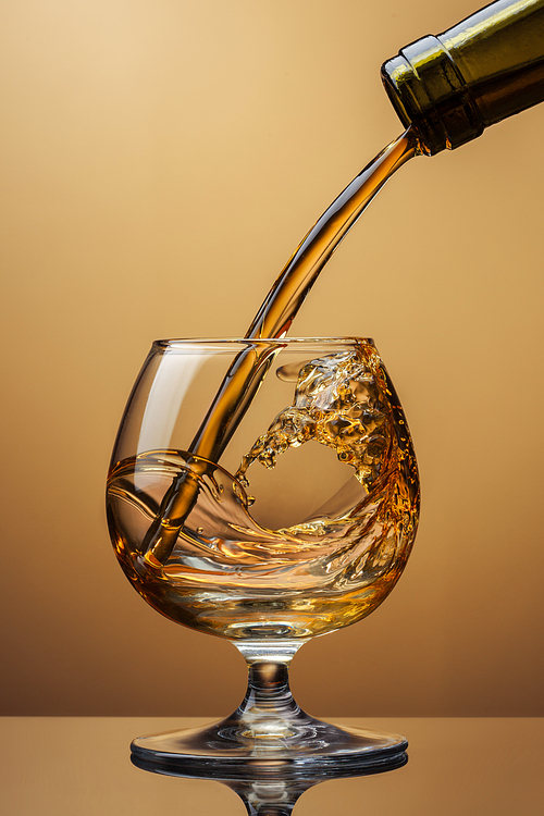 Cognac pouring from bottle into glass with splash on brown background