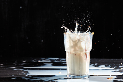 Splashing of milk from the glass with oat muesli and spilled milk on a black wooden table with place for text.