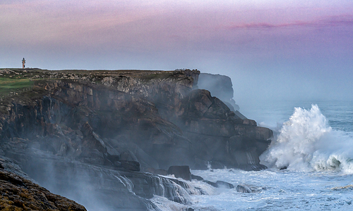 huge storm waves crash onto the Cape Ajo in Spain with the lighthouse on the cliffs above at sunrise