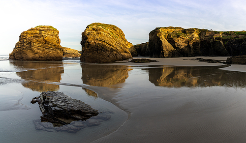 A sandy beach with tidal pools and jagged broken cliffs behind in warm evening light