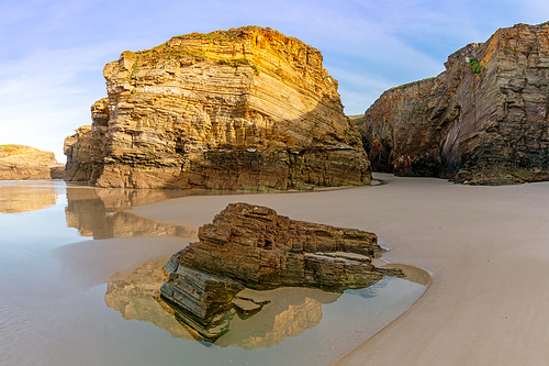 A sandy beach with tidal pools and jagged broken cliffs behind in warm evening light