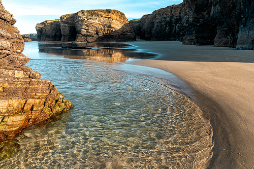 A sandy beach with tidal pools and jagged broken cliffs behind in warm evening light