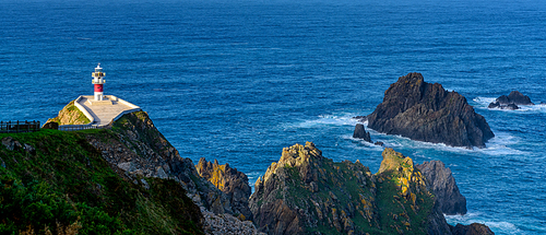 A panorama of the Cabo Ortegal lighthouse in Galicia with green cliffs and sunlight and deep blue ocean