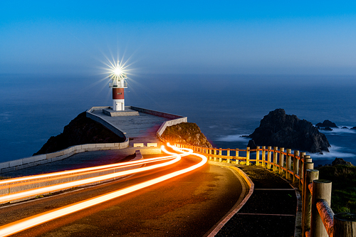 Bright trailing car lights lead to Cabo Ortegal lighthouse at night