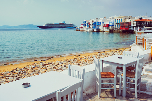 Tourist greek scene - restaurant cafe table on quay promenade with cruise liner and Aegean sea and Little Venice houses in background on beautiful summer day. Chora, Mykonos island, Greece