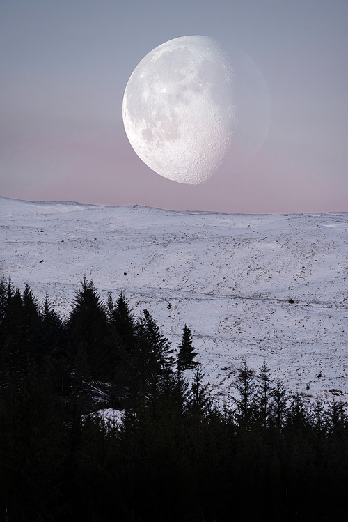 Beautiful composite image of pastel Winter landscape image of giant moon setting behind mountain peak in Scottish Highlands at sunrise