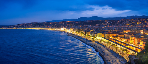 Scenic panorama of Nice, France in evening blue hour. Mediterranean Sea waves surging on coast, people relaxing on beach, lights illumination on colorful houses.