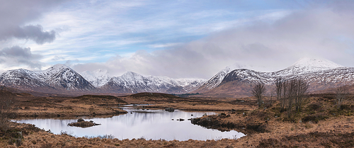 Stunning Winter panorama landscape image of mountain range viewed from Loch Ba in Scottish Highlands with dramatic clouds overhead