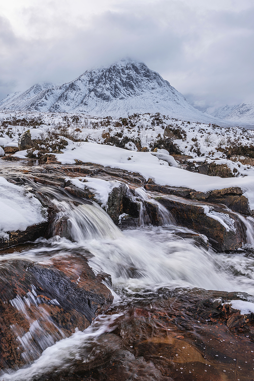 Beautiful Winter landscape image of River Etive in foreground with iconic snowcapped Stob Dearg Buachaille Etive Mor mountain in the background