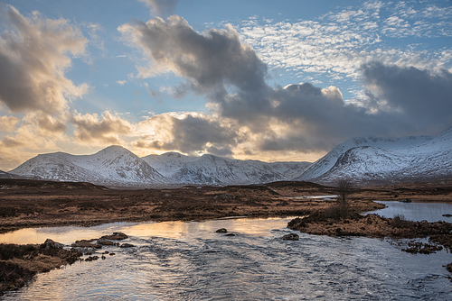 Stunning Winter panorama landscape image of mountain range viewed from Loch Ba in Scottish Highlands with dramatic clouds overhead