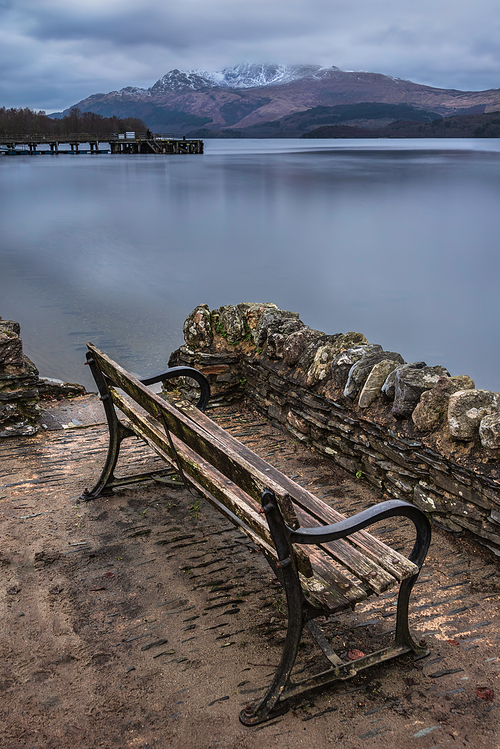 Stunning landscape image of Loch Lomond and snowcapped mountain range in distance viewed from small village of Luss