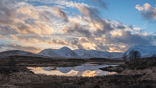 Stunning Winter panorama landscape image of mountain range viewed from Loch Ba in Scottish Highlands with dramatic clouds overhead