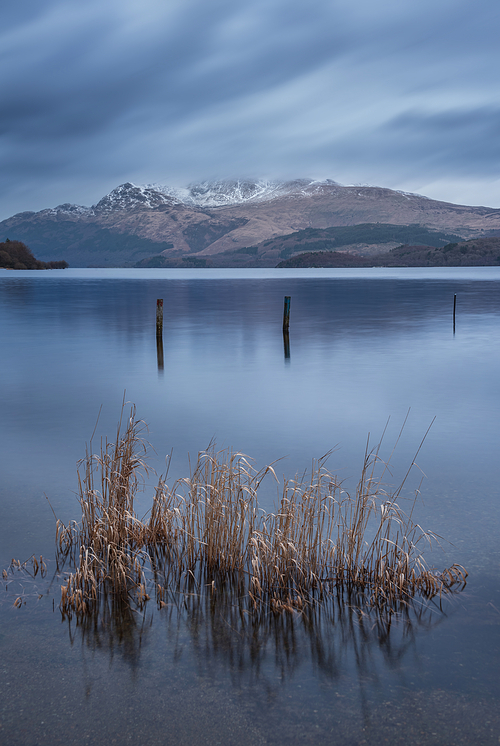 Stunning landscape image of Loch Lomond and snowcapped mountain range in distance viewed from small village of Luss