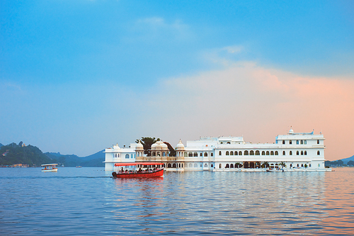 Romantic luxury India travel tourism - tourist boat in front of Lake Palace (Jag Niwas) complex on Lake Pichola on sunset with dramatic sky, Udaipur, Rajasthan, India