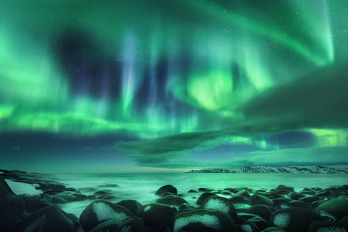 Aurora borealis over ocean. Northern lights in Teriberka, Russia. Starry sky with polar lights and clouds. Night winter landscape with bright aurora, stars, sea, snowy stones in blurred water. Travel