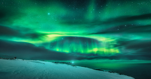 Aurora borealis over ocean. Northern lights in Teriberka, Russia. Starry sky with polar lights and clouds. Night winter landscape with aurora, sea with blurred water, snowy hills, stars. Travel